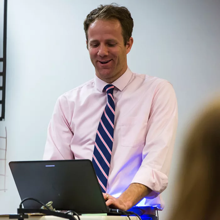 Teacher looks down at laptop, standing and smiling, at front of room
