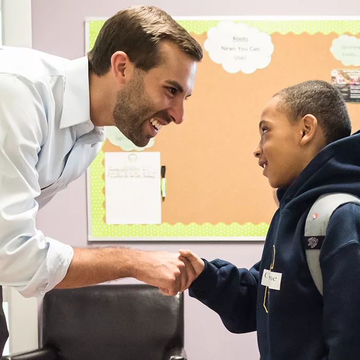 Teacher leans down and shakes child's hand, both smiling
